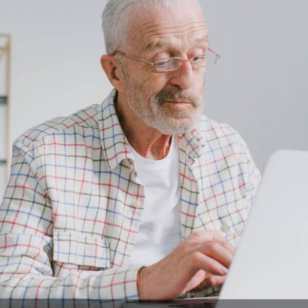 Na zdjęciu mężczyzna w podeszłym wieku w okularach korekcyjnych, patrzący w laptop. Ubrany w biały podkoszulek, rozpiętą, jasną koszule w kartkę czerwono-niebiesko-zieloną. W tle widać białą ścianę.