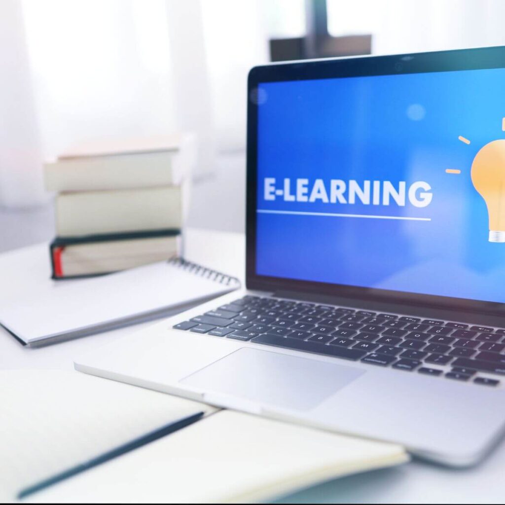 Na zdjęciu znajduje się laptop, którego ekran wyświetla napis „E-LEARNING” oraz grafikę żarówki — klasyczny symbol pomysłowości i nauki. Obok komputera leżą zeszyty, książki oraz notatki.